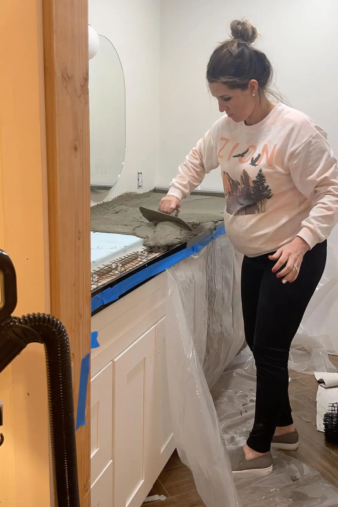 A woman spreading wet concrete over a countertop mold using a trowel. The workspace is lined with protective plastic sheeting, and blue painter's tape secures the edges. An oval mirror is mounted on the wall above.